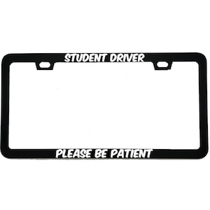 May include: Black license plate frame with white text that reads "STUDENT DRIVER" at the top and "PLEASE BE PATIENT" at the bottom. The frame has a rectangular shape with rounded corners and mounting holes.