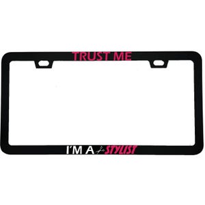 May include: Black license plate frame with the text "TRUST ME" in pink at the top and "I'M A STYLIST" at the bottom. The text is in a bold, sans-serif font. A pair of scissors is used as the "A" in stylist.