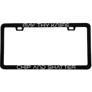 May include: Black license plate frame with the text "MAY THY KNIFE" at the top and "CHIP AND SHATTER" at the bottom. The frame has a rectangular shape with rounded corners and mounting holes.