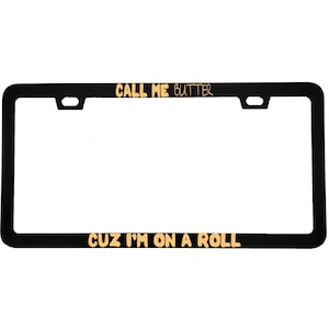 May include: Black license plate frame with gold lettering that reads "CALL ME BUTTER" at the top and "CUZ I'M ON A ROLL" at the bottom. The frame has a rectangular shape with rounded corners.