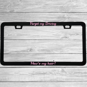 May include: Black license plate frame with the text "Forget my Driving" and "How's my hair?" in pink script. The frame has a rectangular shape with cutouts for mounting. The background is a light gray wood grain.