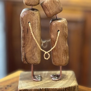 Könnte beinhalten: Eine Holzskulptur, die zwei durch ein Seil verbundene Figuren darstellt, auf einem Holzsockel. Die Figuren bestehen aus rechteckigen Holzblöcken mit Beinen und Köpfen aus Kupferdraht. Die Skulptur hat eine rustikale, handgefertigte Ästhetik.