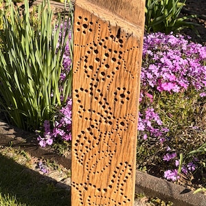 Könnte beinhalten: Ein hoher, rechteckiger Gartenpfahl aus Holz mit natürlicher Oberfläche. Der Pfahl hat dekorative, gebohrte Muster. Die Oberseite ist abgewinkelt. Der Pfahl steht in einem Garten mit grünem Gras und lila Blumen.