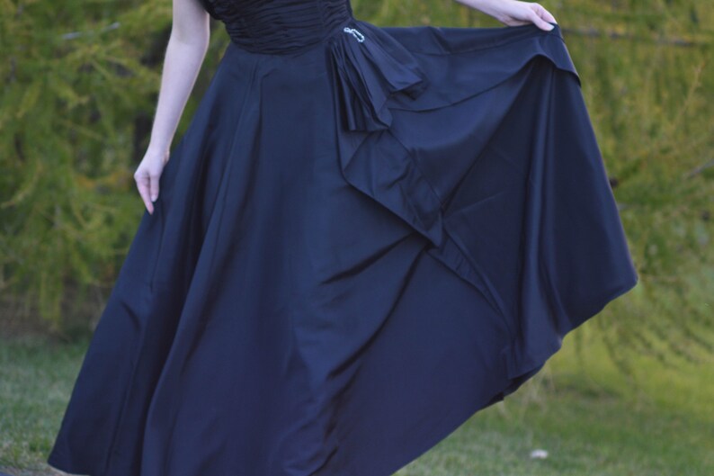 Pu&ograve; includere: Un abito maxi nero con un corpetto drappeggiato e una gonna fluida. La gonna ha un design a strati con un dettaglio a balze.