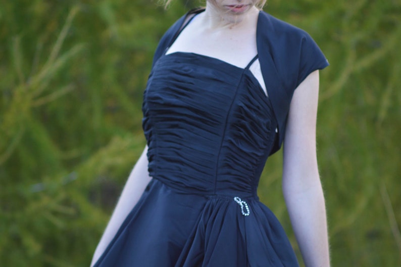Pu&ograve; includere: Una donna indossa un abito blu scuro con un corpetto a balze e una giacca bolero abbinata. L'abito ha un corpetto aderente e una gonna fluida.