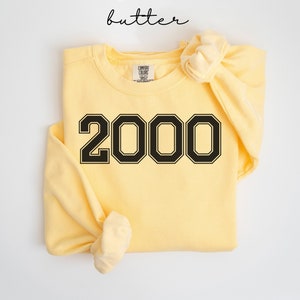 May include: A butter-colored sweatshirt with the number "2000" in a black, collegiate-style font. The word "butter" is written in a cursive font at the top. The sweatshirt is folded with the sleeves tied in a knot.