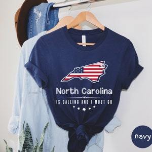 May include: Navy blue t-shirt with a white outline of the state of North Carolina with an American flag design. The text "North Carolina is calling and I must go" is printed below the state outline.