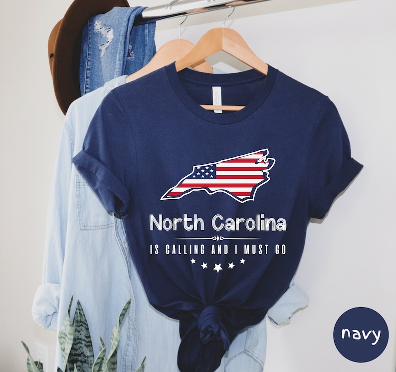 May include: Navy blue t-shirt with a white outline of the state of North Carolina with an American flag design. The text "North Carolina is calling and I must go" is printed below the state outline.