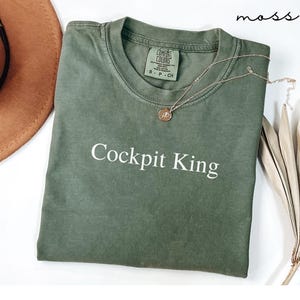 May include: An olive green t-shirt with the text "Cockpit King" in white. A gold necklace with a pendant rests on the shirt. A brown hat and dried plant are in the background. The word "moss" is written in the top right corner.