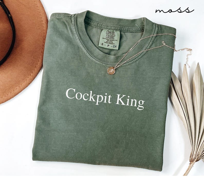 May include: An olive green t-shirt with the text "Cockpit King" in white. A gold necklace with a pendant rests on the shirt. A brown hat and dried plant are in the background. The word "moss" is written in the top right corner.