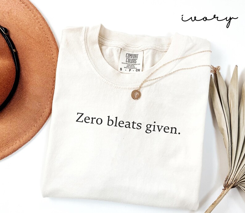 May include: Ivory t-shirt with the text "Zero bleats given." printed in black. The shirt is laying flat on a white surface with a brown hat and a dried plant in the background.