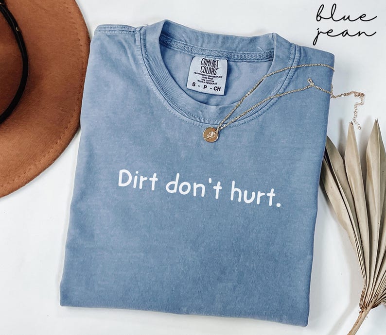 以下が含まれることがあります： 白字で「Dirt don't hurt」と書かれたブルーデニムのComfort Colors Tシャツ。ペンダント付きのゴールドネックレスがシャツの上にかけられています。シャツは折りたたまれ、白い表面に置かれています。