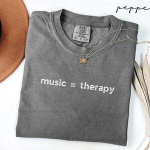 May include: Gray t-shirt with the text "music = therapy" embroidered in white. The shirt is made of a soft, comfortable fabric and has a relaxed fit.
