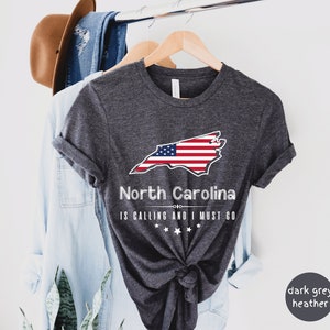 May include: Dark grey heather t-shirt with a white outline of the state of North Carolina with an American flag design. The text "North Carolina is calling and I must go" is printed below the state outline.