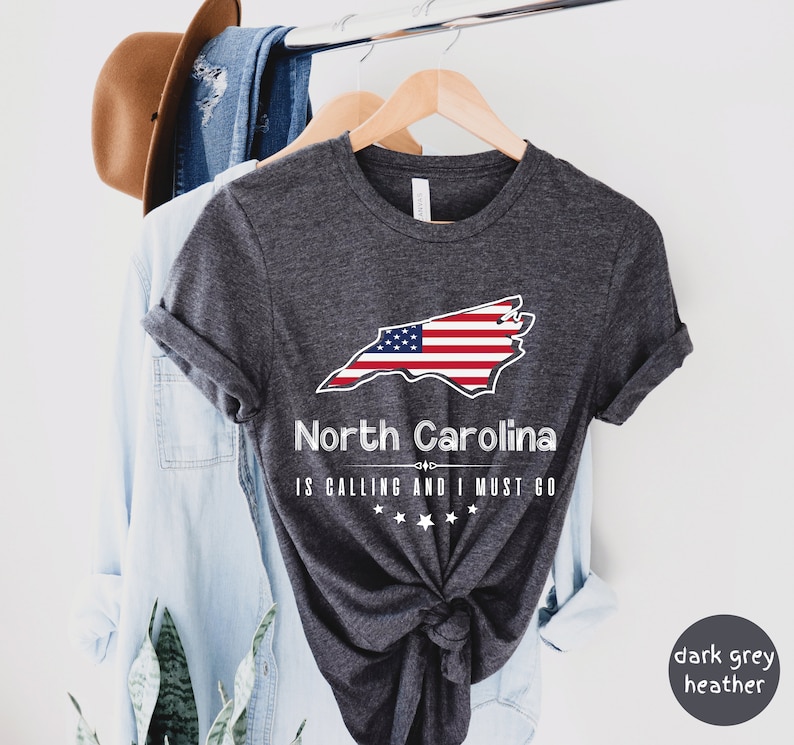 May include: Dark grey heather t-shirt with a white outline of the state of North Carolina with an American flag design. The text "North Carolina is calling and I must go" is printed below the state outline.