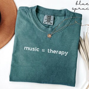 May include: A teal blue t-shirt with the text "music = therapy" in white embroidery. The shirt is made of a soft, comfortable fabric and has a relaxed fit.