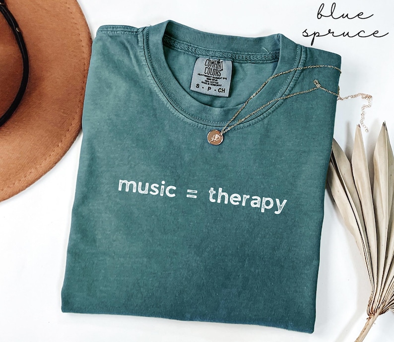 May include: A teal blue t-shirt with the text "music = therapy" in white embroidery. The shirt is made of a soft, comfortable fabric and has a relaxed fit.