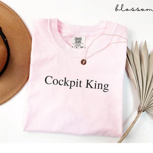 May include: A light pink t-shirt with the text "Cockpit King" in black. A gold necklace with a pendant is draped over the shirt. The shirt is folded on a white surface, with a brown hat and a dried palm leaf visible.