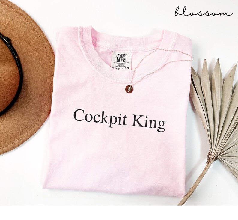 May include: A light pink t-shirt with the text "Cockpit King" in black. A gold necklace with a pendant is draped over the shirt. The shirt is folded on a white surface, with a brown hat and a dried palm leaf visible.