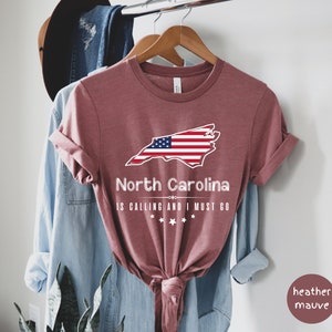 May include: Heather mauve t-shirt with a graphic of the state of North Carolina with the American flag superimposed on it. The text "North Carolina is calling and I must go" is printed below the state outline.