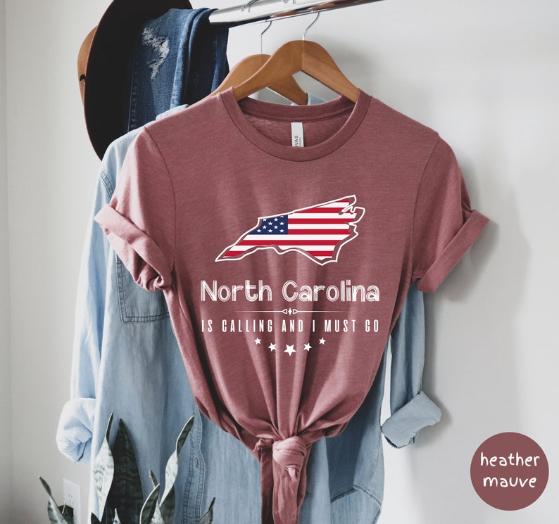 May include: Heather mauve t-shirt with a graphic of the state of North Carolina with the American flag superimposed on it. The text "North Carolina is calling and I must go" is printed below the state outline.