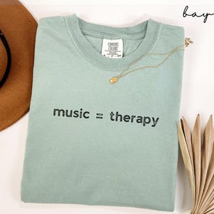 May include: A light green t-shirt with the text "music = therapy" printed in black. The shirt is folded and laid flat on a white surface.