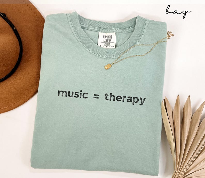 May include: A light green t-shirt with the text "music = therapy" printed in black. The shirt is folded and laid flat on a white surface.
