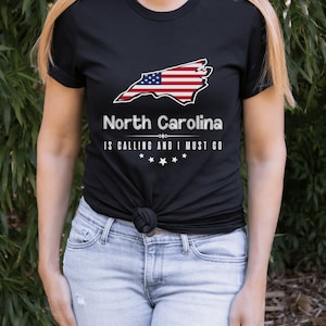 May include: Black t-shirt with a white outline of the state of North Carolina with an American flag design. The text "North Carolina is calling and I must go" is printed below the state outline.