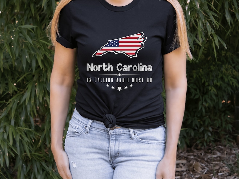 May include: Black t-shirt with a white outline of the state of North Carolina with an American flag design. The text "North Carolina is calling and I must go" is printed below the state outline.