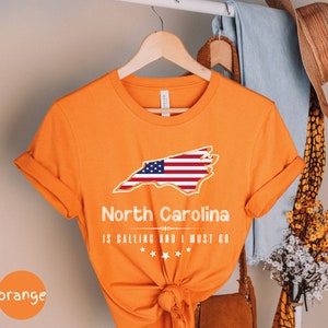 May include: Orange t-shirt with a white outline of the state of North Carolina with an American flag inside. The text "North Carolina is calling and I must go" is printed below the state outline.