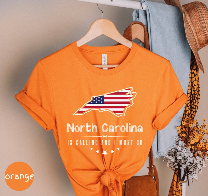 May include: Orange t-shirt with a white outline of the state of North Carolina with an American flag inside. The text "North Carolina is calling and I must go" is printed below the state outline.