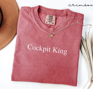 May include: Crimson-colored t-shirt with the text "Cockpit King" in white. The shirt has a crew neck and a small tag on the collar. A silver necklace with a pendant is draped across the shirt. The shirt is folded on a white surface.