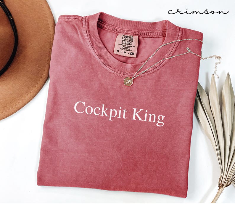 May include: Crimson-colored t-shirt with the text "Cockpit King" in white. The shirt has a crew neck and a small tag on the collar. A silver necklace with a pendant is draped across the shirt. The shirt is folded on a white surface.