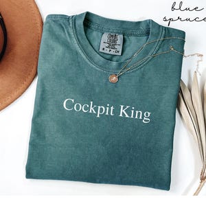 May include: Blue spruce-colored t-shirt with the text "Cockpit King" in white. The shirt has a crew neck and a small tag on the collar. A gold necklace with a pendant is draped across the shirt. A brown hat and dried leaves are in the background.
