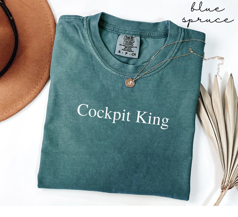 May include: Blue spruce-colored t-shirt with the text "Cockpit King" in white. The shirt has a crew neck and a small tag on the collar. A gold necklace with a pendant is draped across the shirt. A brown hat and dried leaves are in the background.