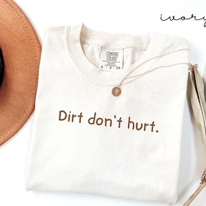 以下が含まれることがあります： 「Dirt don't hurt」という茶色の文字がプリントされたアイボリー色のTシャツ。ペンダント付きのゴールドネックレスがシャツの上にあります。茶色の帽子と乾燥した植物が背景に見えます。