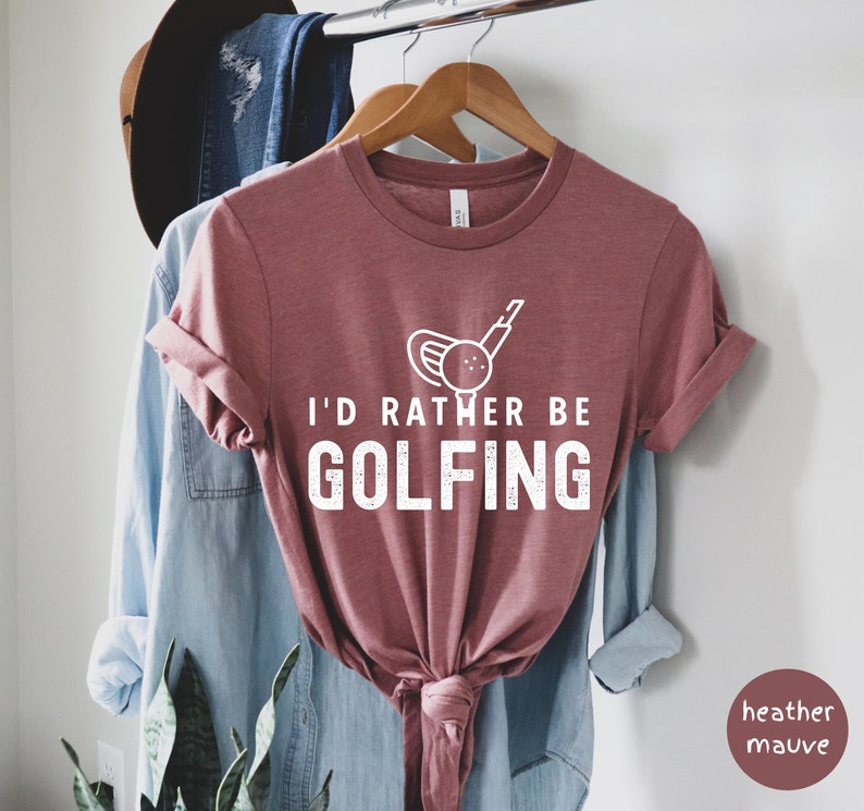 May include: A heather mauve t-shirt with a white graphic of a golf club and ball. The text "I'd rather be golfing" is printed in white on the shirt.