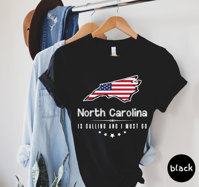 May include: Black t-shirt with a white outline of the state of North Carolina with an American flag design. The text "North Carolina is calling and I must go" is printed below the state outline.