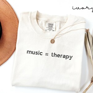 May include: Ivory t-shirt with the text "music = therapy" printed in black. The shirt is laying flat on a white surface with a brown hat and a dried palm leaf.