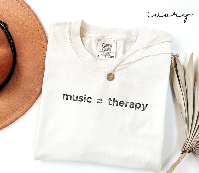 May include: Ivory t-shirt with the text "music = therapy" printed in black. The shirt is laying flat on a white surface with a brown hat and a dried palm leaf.
