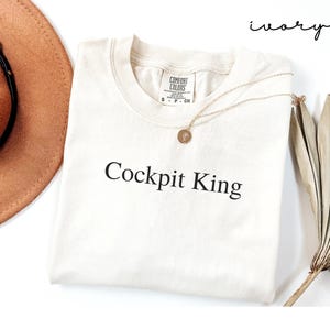 May include: Ivory-colored t-shirt with the text "Cockpit King" in black. The shirt has a crew neck and short sleeves. A gold necklace with a circular pendant is draped on the shirt. A brown hat and dried plant are in the background.