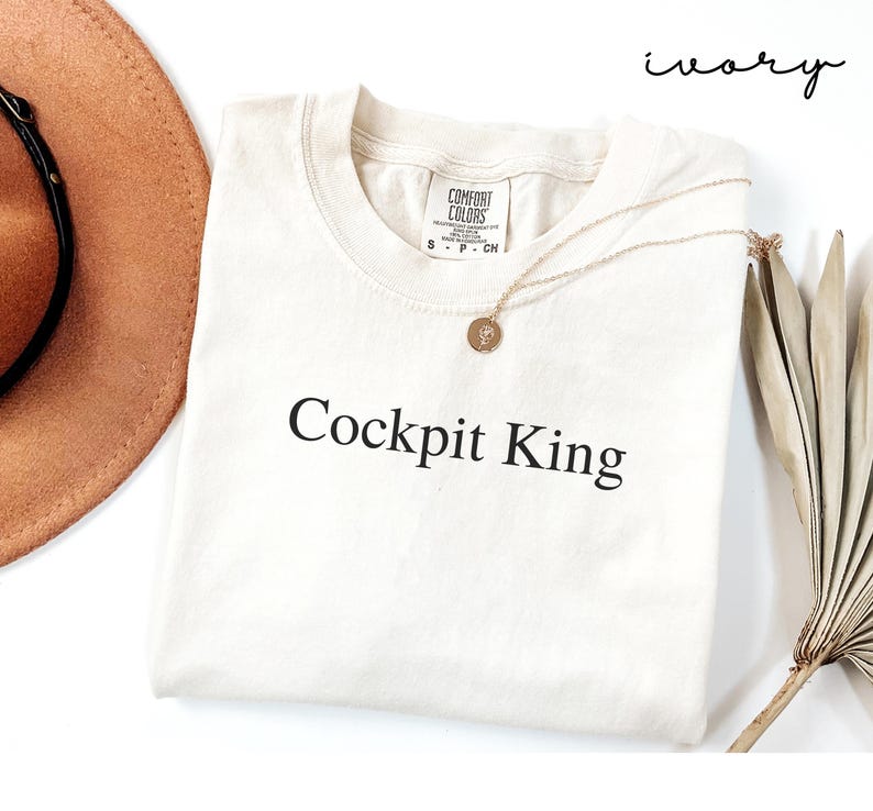 May include: Ivory-colored t-shirt with the text "Cockpit King" in black. The shirt has a crew neck and short sleeves. A gold necklace with a circular pendant is draped on the shirt. A brown hat and dried plant are in the background.