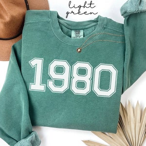 May include: Light green sweatshirt with the year 1980 printed in white on the front.