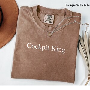 May include: Espresso-colored t-shirt with the text "Cockpit King" in white. The shirt has a crew neck and a Comfort Colors tag. A silver necklace with a pendant is draped across the shirt. A brown hat and dried leaves are in the background.