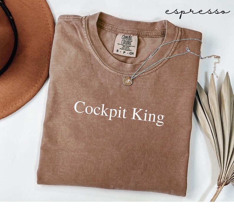 May include: Espresso-colored t-shirt with the text "Cockpit King" in white. The shirt has a crew neck and a Comfort Colors tag. A silver necklace with a pendant is draped across the shirt. A brown hat and dried leaves are in the background.