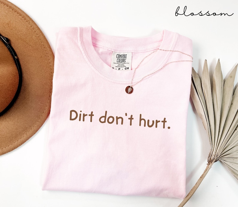 以下が含まれることがあります： 「Dirt don't hurt」という茶色の文字がプリントされた、薄いピンク色のTシャツ。ペンダント付きのゴールドネックレスがシャツの上にあります。茶色の帽子と乾燥した植物が背景にあります。