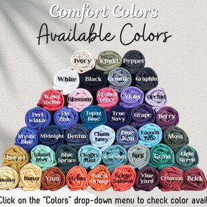 May include: A variety of rolled fabric swatches in different colors, including ivory, khaki, pepper, white, black, granite, graphite, water melon, blossom, crunch berry, violet, orchid, peri winkle, flo blue, topaz blue, true navy, grape, berry, mystic blue, midnight, denim, cham bray, blue jean, lagoon blue, moss, mustard, island reef, blue spruce, chalky mint, seafoam, bay, grass, light green, banana, butter, yam, melon, burnt orange, bright salmon, vine yard, crimson, and brick. The text "Comfort Colors Available Colors" is at the top of the image. The text "Note: Click on the "Colors" drop-down menu to check color availability" is at the bottom of the image.