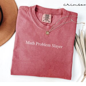 May include: Crimson-colored t-shirt with the text "Math Problem Slayer" in white. A gold-colored necklace with a pendant is draped over the shirt. The shirt is made of a soft material and has a crew neck.