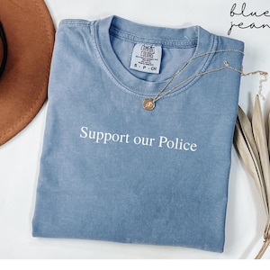 May include: A blue jean Comfort Colors t-shirt with the text "Support our Police" in white. A gold necklace with a pendant is draped across the shirt. The shirt is folded and placed on a white surface.