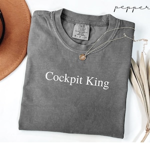 May include: A gray Comfort Colors t-shirt with the text "Cockpit King" in white. The shirt has a crew neck and short sleeves. A gold necklace with a pendant is draped on the shirt. The word "pepper" is written in cursive in the upper right corner.